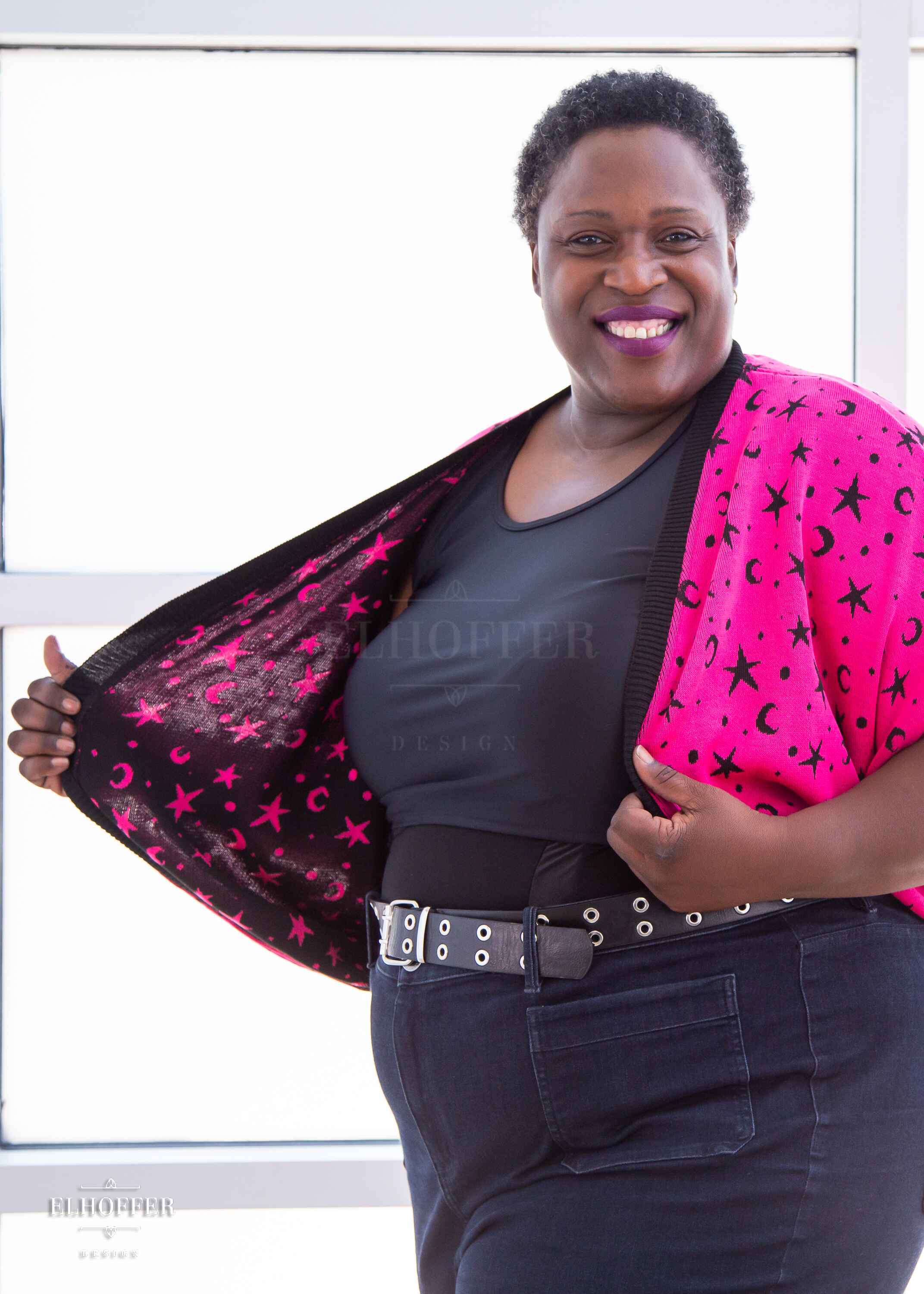 Adalgiza, a medium dark skinned 4xl model with short dark tight curly hair, is smiling while wearing an open front dolman with 3/4 sleeves. The main body of the dolman is bright pink with a black star and moon pattern knit throughout.  The edges and cuffs are ribbed in black.