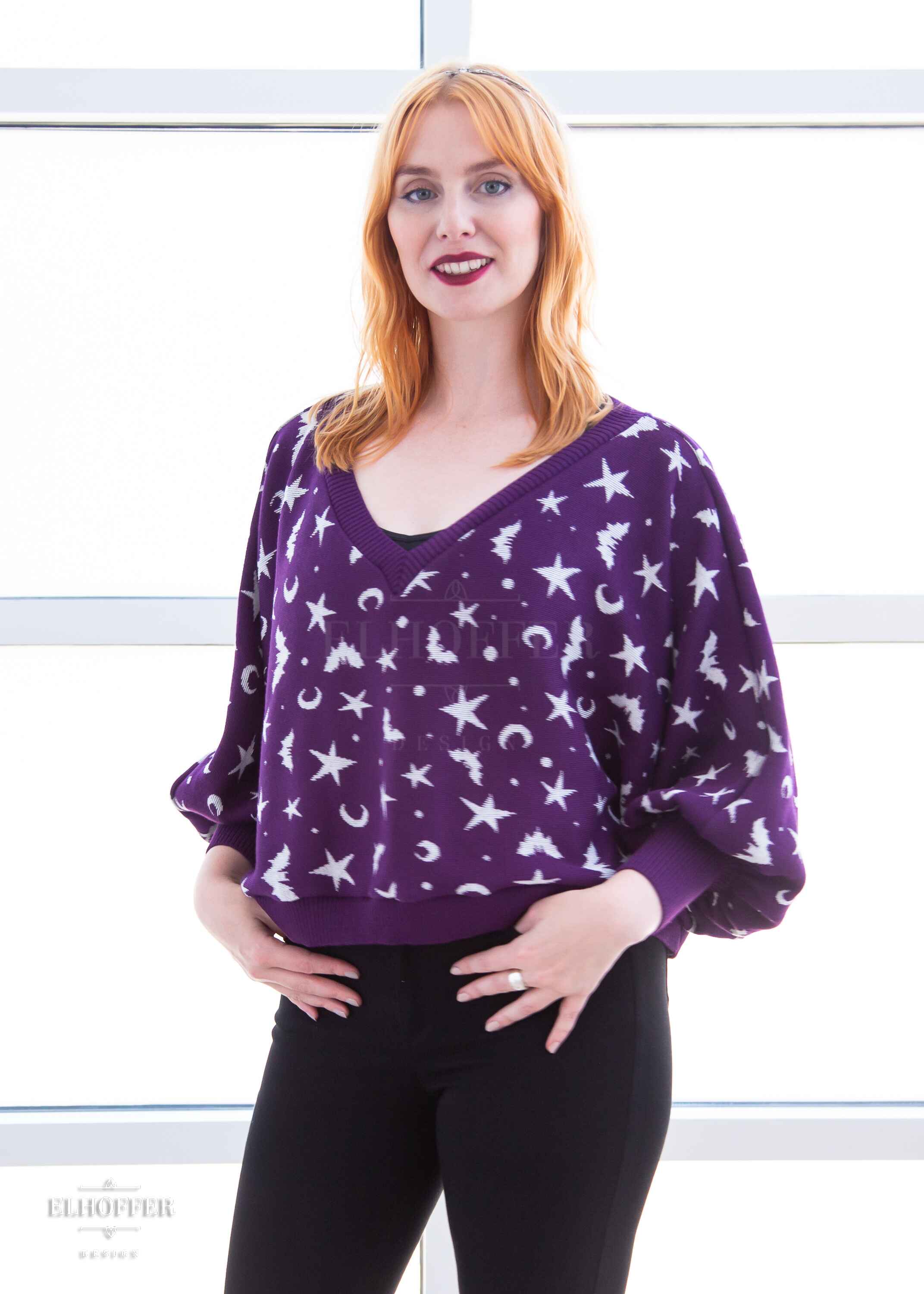 Harley, a fair skinned S model with shoulder length strawberry blonde hair, is smiling while wearing an oversized v neck cropped sweater with batwing sleeves that gather at the wrist. The main body of the sweater is purple with a white bat, star, moon, and dot pattern repeated throughout.