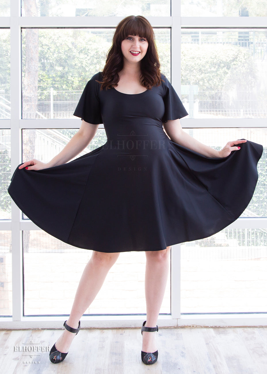 Dayna, a fair skinned medium model with long dark brown hair and bangs, is wearing a knee-length pullover dress with scoop neck, flutter sleeves, and side pockets in black. She's showing off the amount of fabric in the skirt.