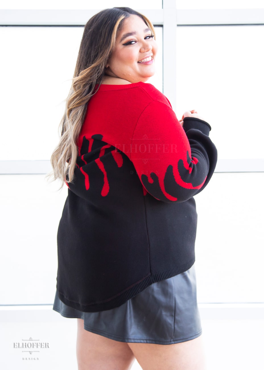 Cori, a sun kissed skinned 2xl model with long balayage hair, is smiling while wearing an oversize sweater with a bright red blood drip design that looks like it's oozing down onto a black sweater that has long billowing sleeves with thumbholes.
