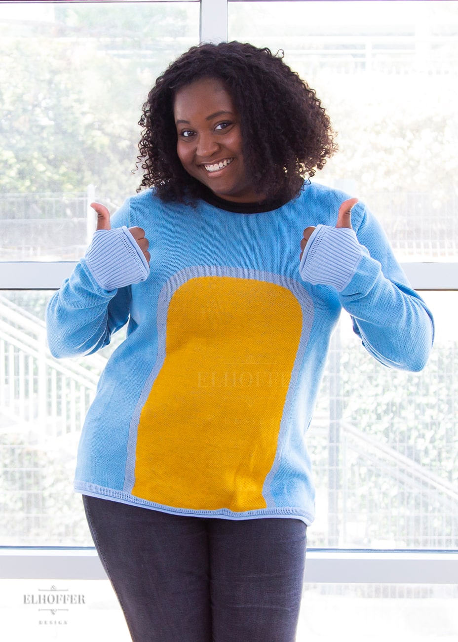 Maydelle, a medium dark skinned XL model with shoulder length curly dark brown hair, is wearing a fitted long sleeve knit sweater with wide crew neck and rolled edges and thumbholes in the sleeve. It is a medium blue with light blue cuffs and detail around the yellow oval in the middle of the sweater. It also has a black neck and black spot in the middle of the back.