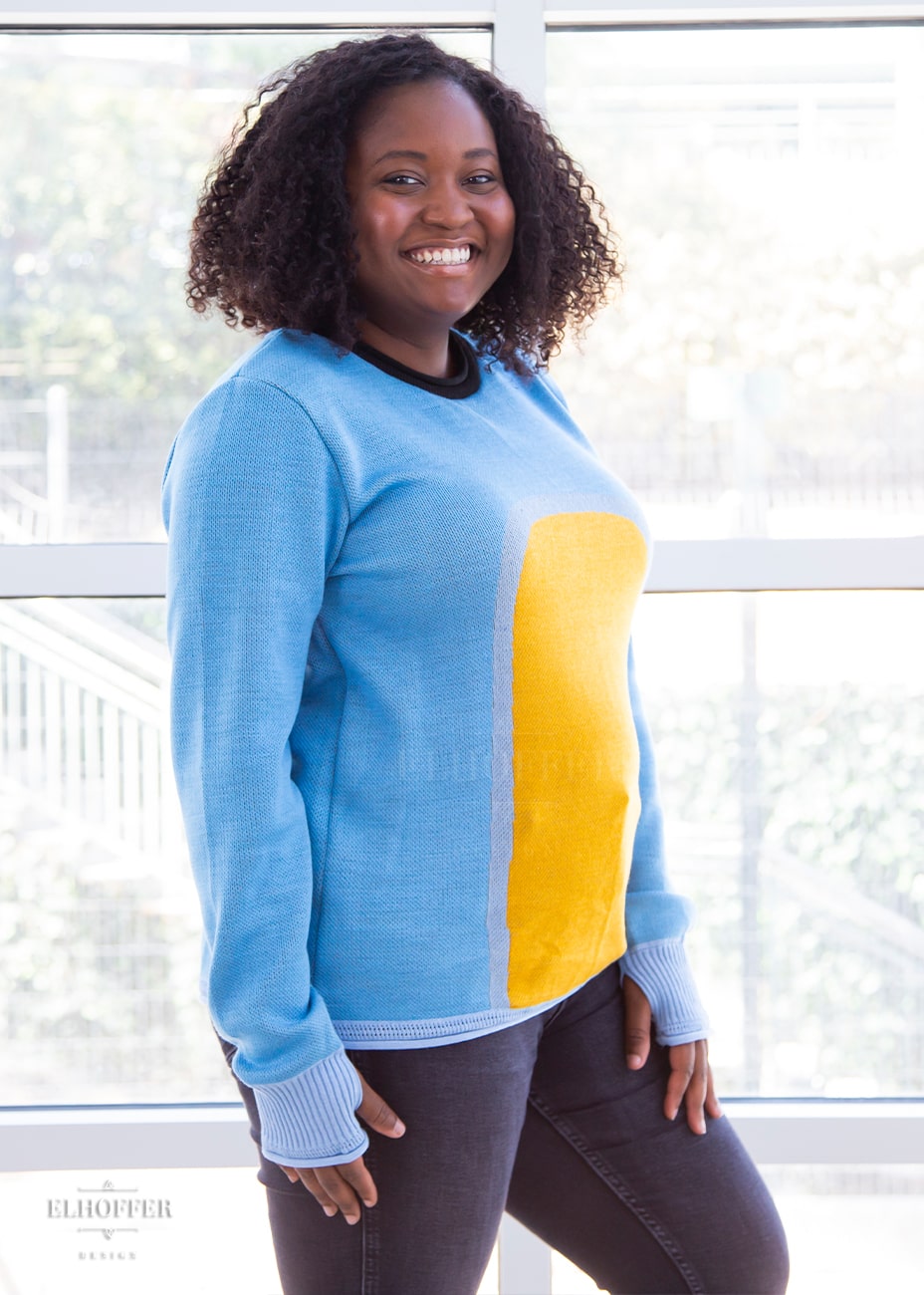 Maydelle, a medium dark skinned XL model with shoulder length curly dark brown hair, is wearing a fitted long sleeve knit sweater with wide crew neck and rolled edges and thumbholes in the sleeve. It is a medium blue with light blue cuffs and detail around the yellow oval in the middle of the sweater. It also has a black neck and black spot in the middle of the back.