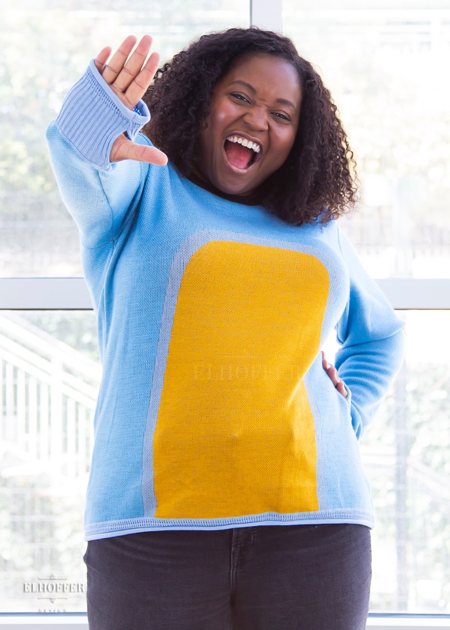 Maydelle, a medium dark skinned XL model with shoulder length curly dark brown hair, is wearing a fitted long sleeve knit sweater with wide crew neck and rolled edges and thumbholes in the sleeve. It is a medium blue with light blue cuffs and detail around the yellow oval in the middle of the sweater. It also has a black neck and black spot in the middle of the back.