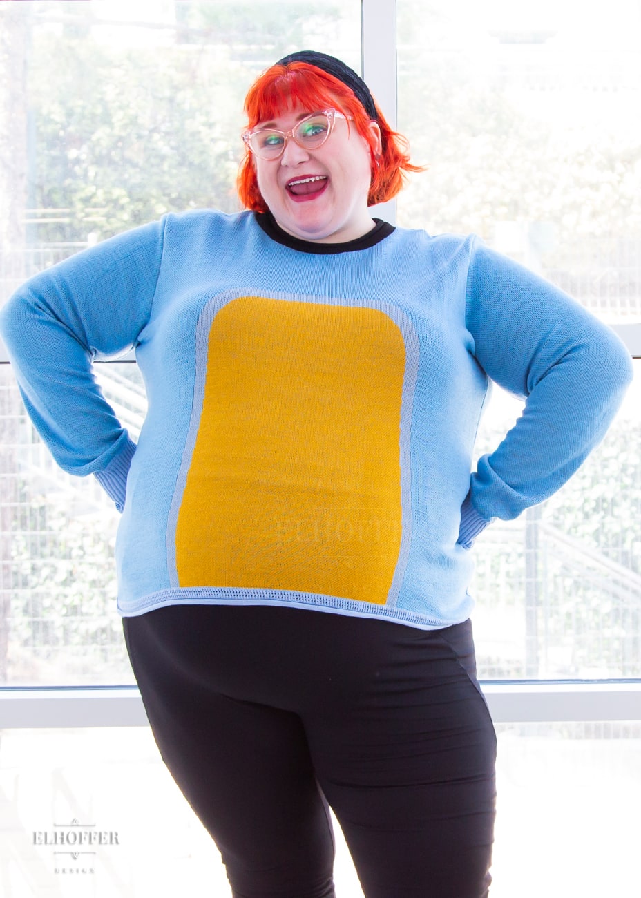 Logan, a fair skinned size 3XL model with short bright orange hair with bangs,  is wearing a fitted long sleeve knit sweater with wide crew neck and rolled edges and thumbholes in the sleeve. It is a medium blue with light blue cuffs and detail around the yellow oval in the middle of the sweater. It also has a black neck and black spot in the middle of the back.