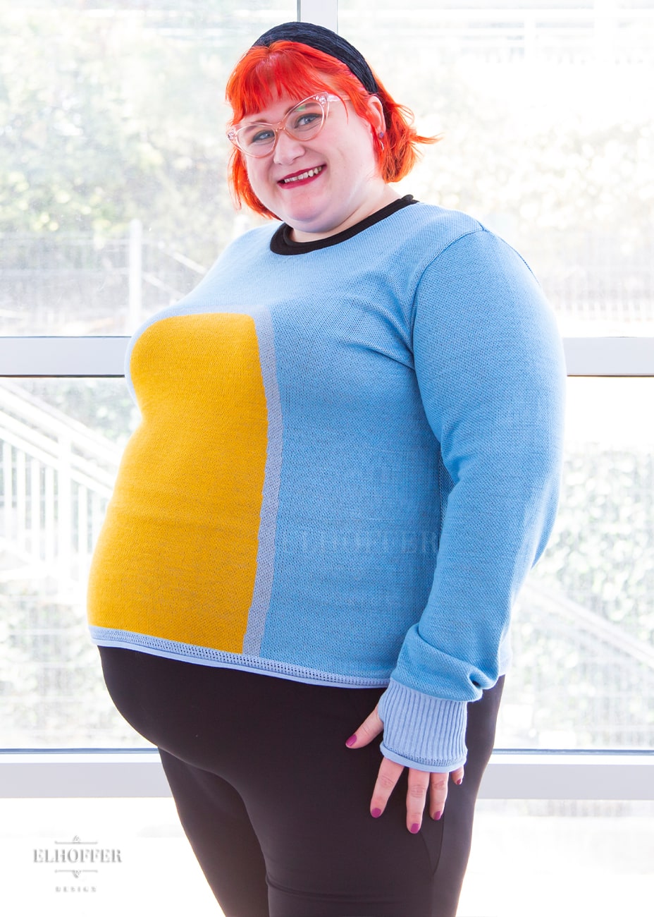 Logan, a fair skinned size 3XL model with short bright orange hair with bangs, is wearing a fitted long sleeve knit sweater with wide crew neck and rolled edges and thumbholes in the sleeve. It is a medium blue with light blue cuffs and detail around the yellow oval in the middle of the sweater. It also has a black neck and black spot in the middle of the back.