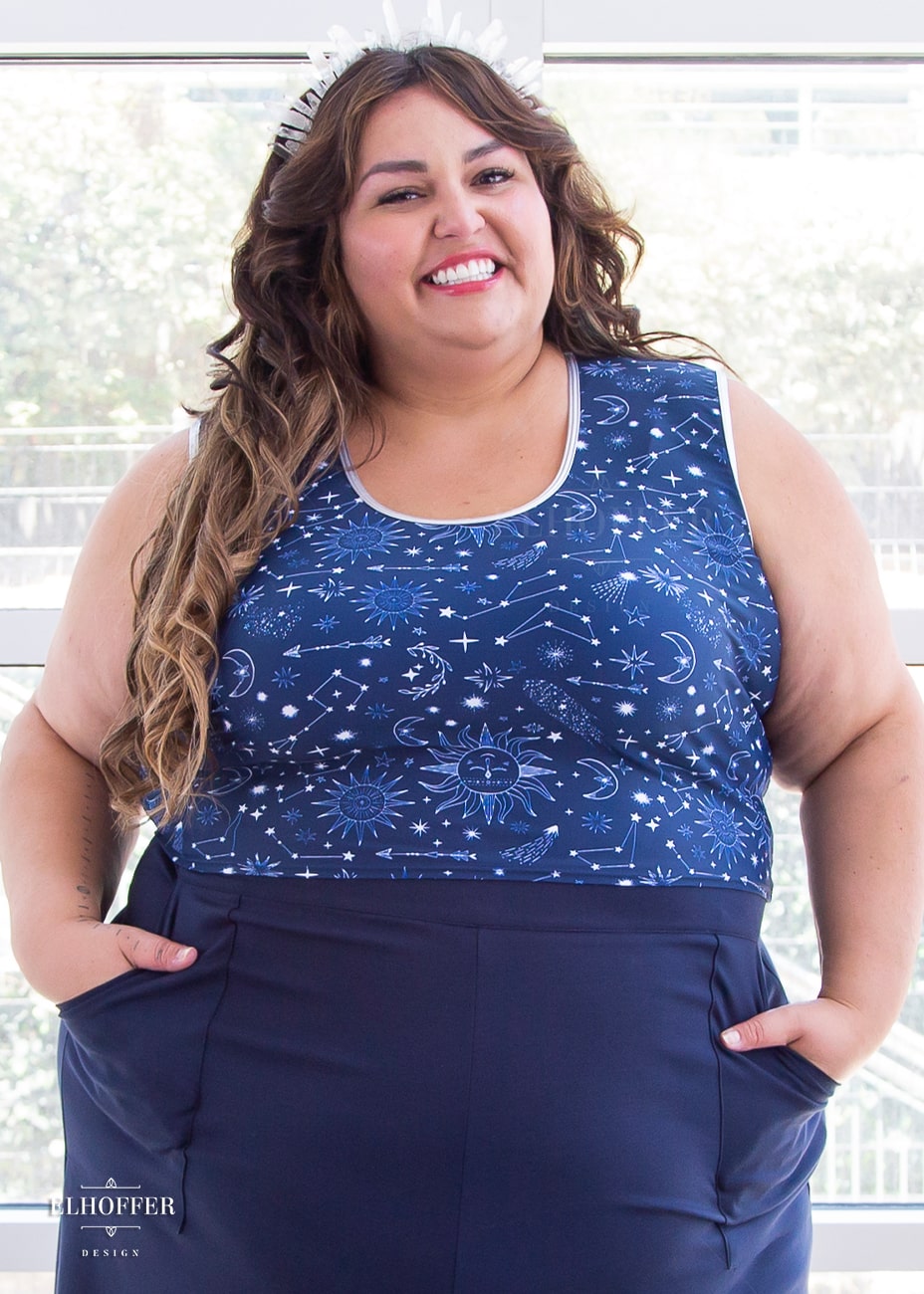 Kristen, a size 3XL olive skinned model with long highlighted brown hair, is wearing a low scoop neckline sleeveless crop top with silver metallic foldover binding at the neck and armholes, wide shoulder straps, fitted throughout the body, and ending at the natural waist in our star crossed lovers print. The Star-Crossed Lovers print is a celestial inspired pattern featuring a navy base and white stars, constellations, sunds, moons, and other small details scattered across the repeated art.