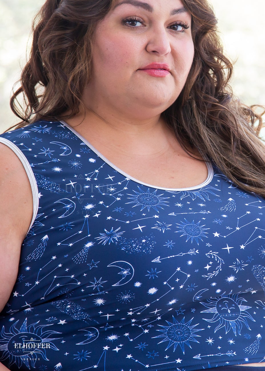 Kristen, a size 3XL olive skinned model with long brown highlighted hair, is wearing a low scoop neckline sleeveless crop top with silver metallic foldover binding at the neck and armholes, wide shoulder straps, fitted throughout the body, and ending at the natural waist in our star crossed lovers print. The Star-Crossed Lovers print is a celestial inspired pattern featuring a navy base and white stars, constellations, sunds, moons, and other small details scattered across the repeated art.