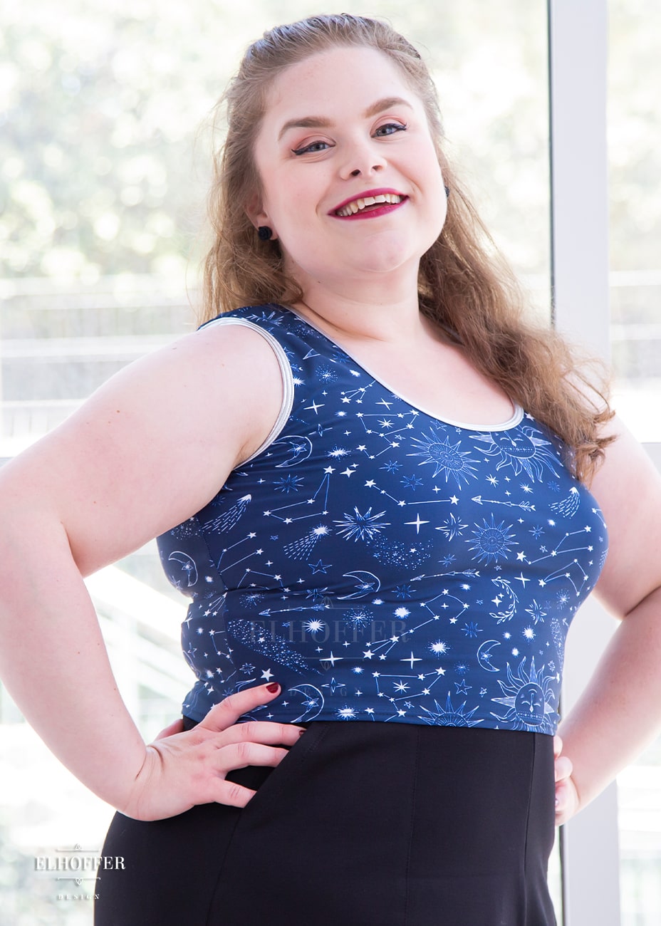 Lucy, a size large fair skinned model with long  dark brown hair, is wearing a low scoop neckline sleeveless crop top with silver metallic foldover binding at the neck and armholes, wide shoulder straps, fitted throughout the body, and ending at the natural waist in our star crossed lovers print. The Star-Crossed Lovers print is a celestial inspired pattern featuring a navy base and white stars, constellations, sunds, moons, and other small details scattered across the repeated art.