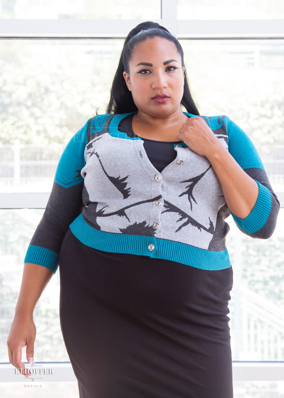 Tas, a medium dark skinned size 2xl model with long dark hair in a pony tail, is wearing a cropped button up knit cardigan with 3/4 sleeves. The cardigan front is mainly a light grey with dark grey decorative lines that give the impression of battle damaged armor, with teal at the shoulders, and the bottom half of the sleeve and back of the cardigan are a dark grey. The neckine, bottom, and cuffs are all teal ribbing.