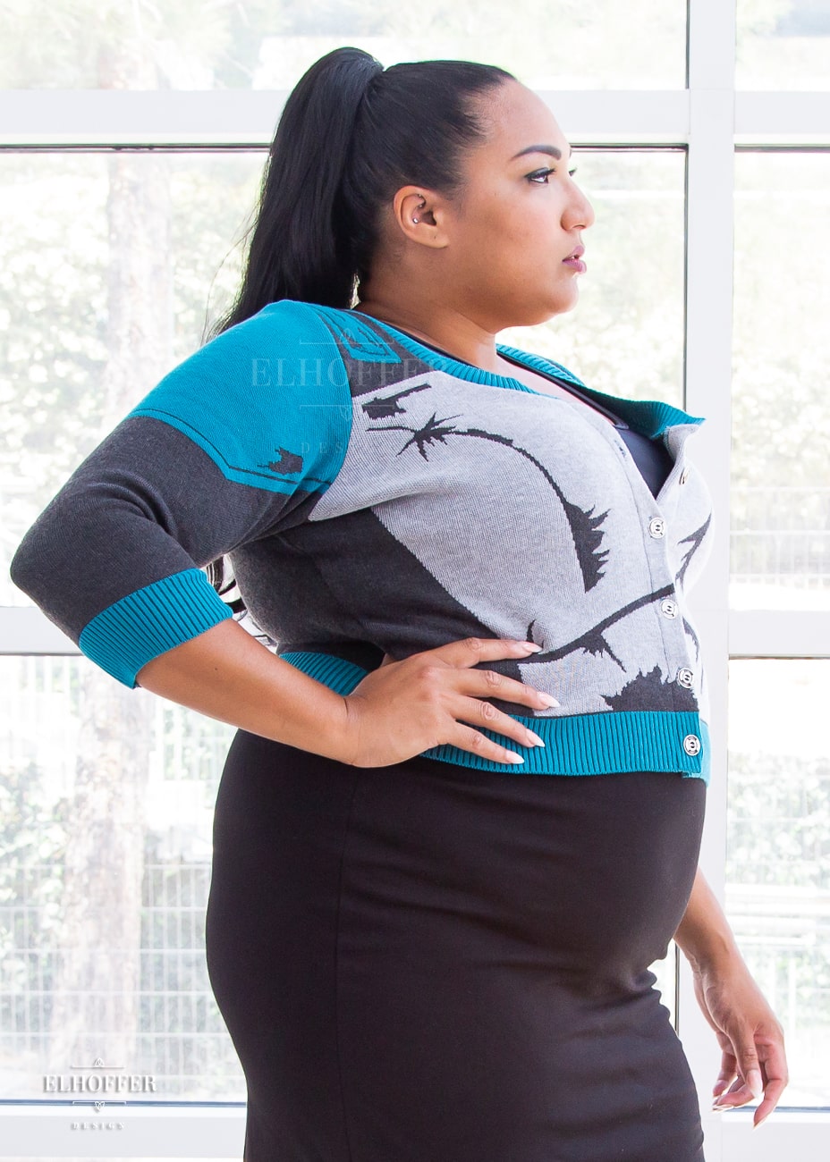 A side view of Tas, a medium dark skinned size 2xl model with long dark hair in a pony tail, wearing a cropped button up knit cardigan with 3/4 sleeves. The cardigan front is mainly a light grey with dark grey decorative lines that give the impression of battle damaged armor, with teal at the shoulders, and the bottom half of the sleeve and back of the cardigan are a dark grey. The neckine, bottom, and cuffs are all teal ribbing.