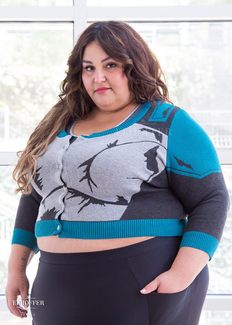 Kristen, a sun kissed skin 4xl model with long wavy brown hair, is wearing a cropped button up knit cardigan with 3/4 sleeves. The cardigan front is mainly a light grey with dark grey decorative lines that give the impression of battle damaged armor, with teal at the shoulders, and the bottom half of the sleeve and back of the cardigan are a dark grey. The neckine, bottom, and cuffs are all teal ribbing.