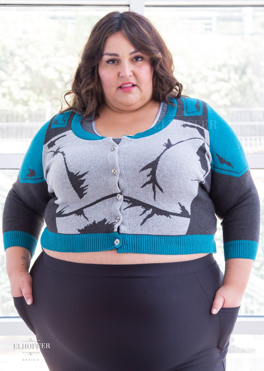Kristen, a sun kissed skin 4xl model with long wavy brown hair, is wearing a cropped button up knit cardigan with 3/4 sleeves. The cardigan front is mainly a light grey with dark grey decorative lines that give the impression of battle damaged armor, with teal at the shoulders, and the bottom half of the sleeve and back of the cardigan are a dark grey. The neckine, bottom, and cuffs are all teal ribbing.