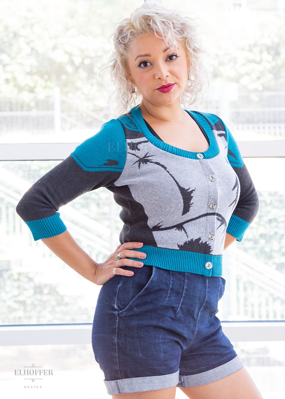 Simone, a size small model with light olive skin and curly platinum hair, is wearing a cropped button up knit cardigan with 3/4 sleeves. The cardigan front is mainly a light grey with dark grey decorative lines that give the impression of battle damaged armor, with teal at the shoulders, and the bottom half of the sleeve and back of the cardigan are a dark grey. The neckine, bottom, and cuffs are all teal ribbing.