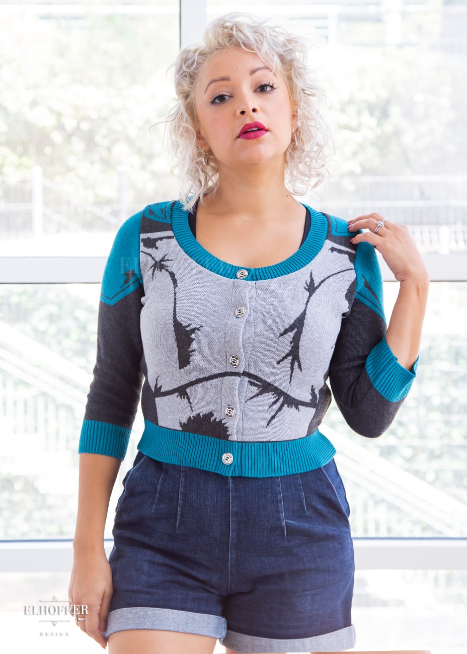 Simone, a size small model with light olive skin and curly platinum hair, is wearing a cropped button up knit cardigan with 3/4 sleeves. The cardigan front is mainly a light grey with dark grey decorative lines that give the impression of battle damaged armor, with teal at the shoulders, and the bottom half of the sleeve and back of the cardigan are a dark grey. The neckine, bottom, and cuffs are all teal ribbing.