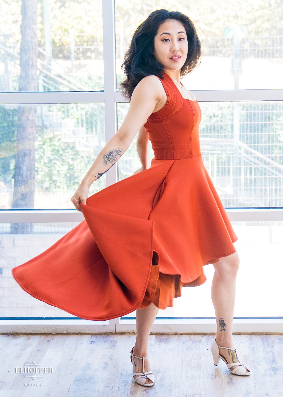 Kate, a size XS olive skinned model with long dark hair, is wearing a rust orange dress with a scoop neck, quilted sleeveless shoulders, with hooks at the back. The skirt is high low, falling to the knees at the front and ankles at the back.