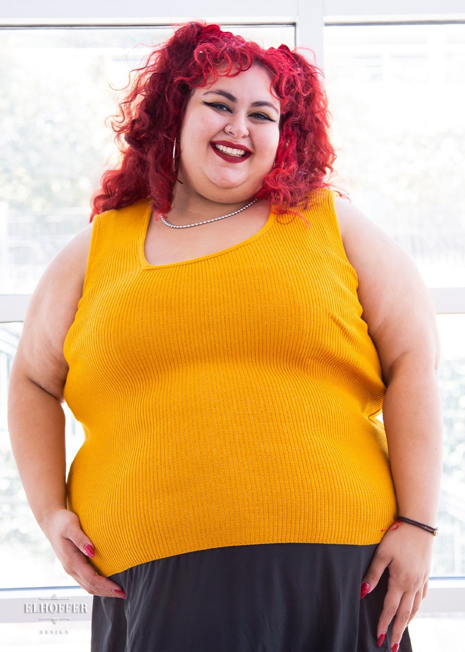 Victoria, an olive skinned size 4XL model with bright red curly hair, is wearing a pullover sleeveless ribbed sweater tank with an asymmetrical neckline in mustard yellow.