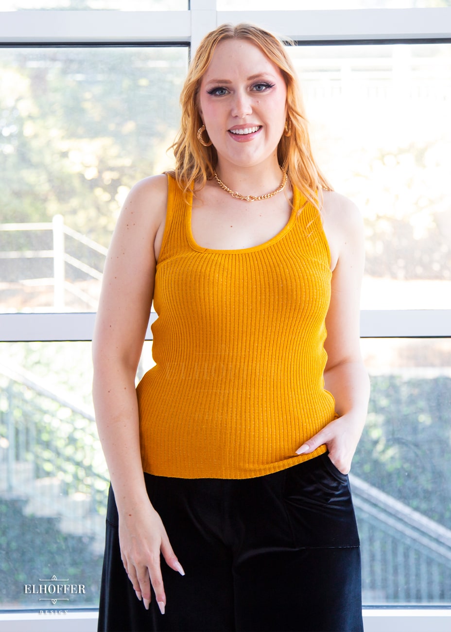 Kelsey, a fair skinned S model with light red hair, is wearing a pullover sleeveless ribbed sweater tank with an asymmetrical neckline in mustard gold.