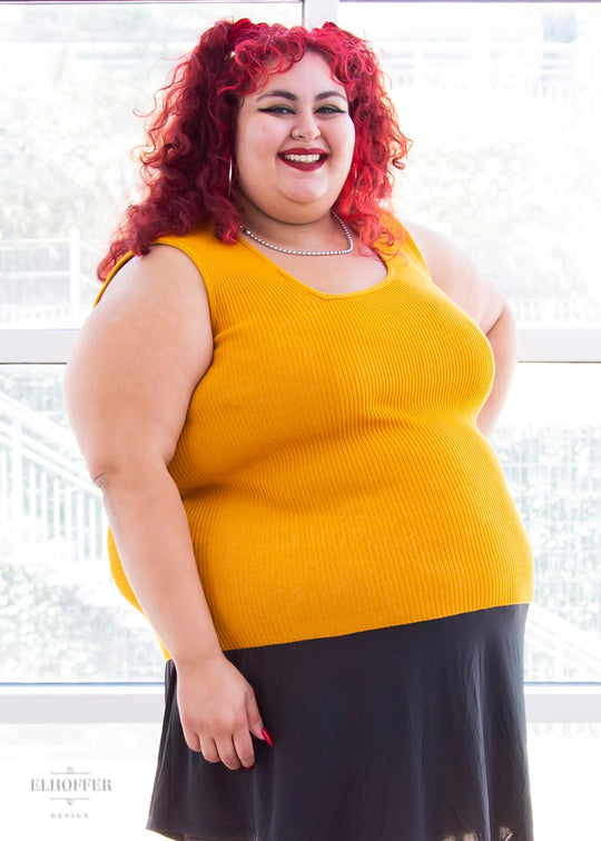 Victoria, an olive skinned size 4XL model with bright red curly hair, is wearing a pullover sleeveless ribbed sweater tank with an asymmetrical neckline in mustard yellow.