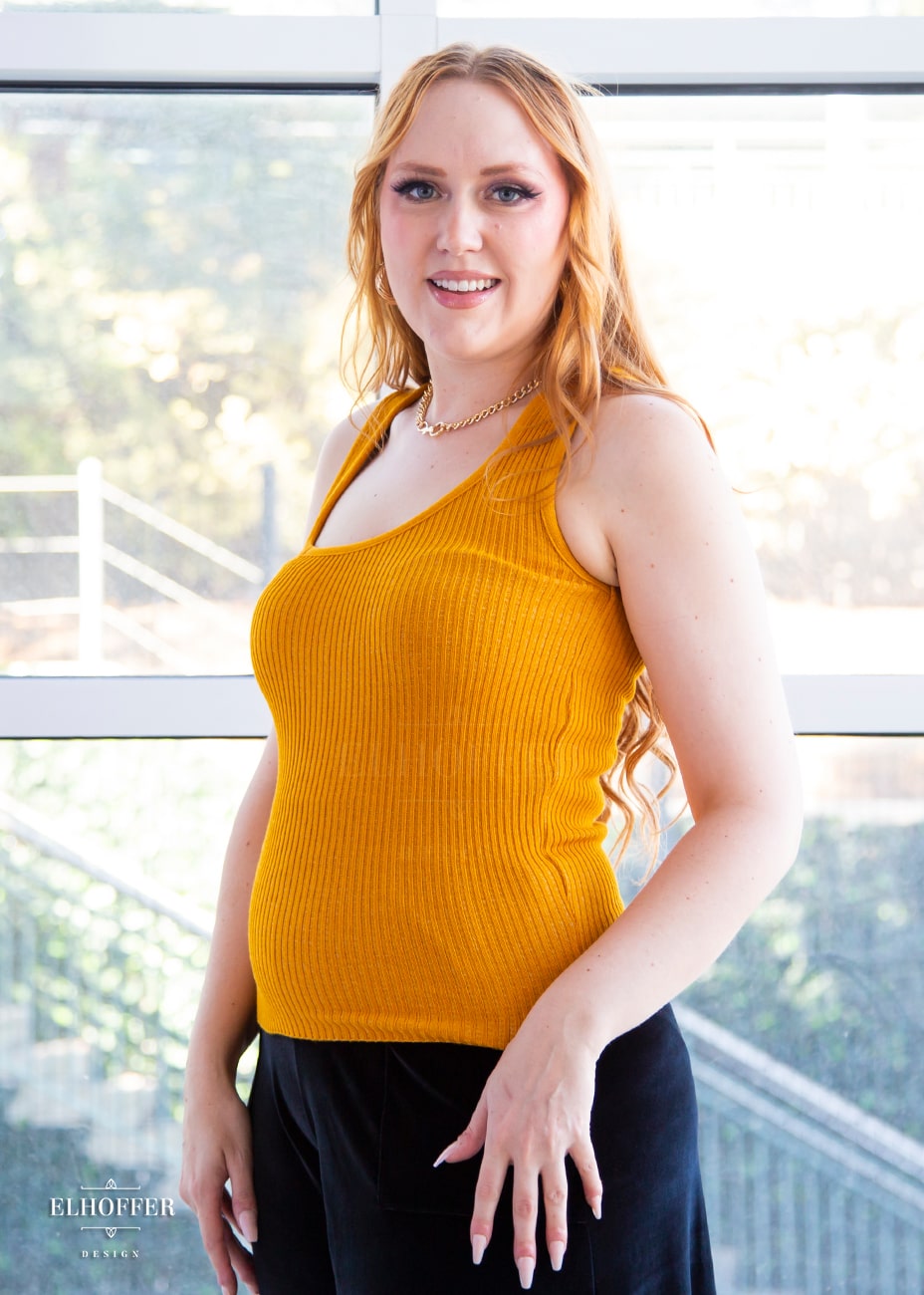 Kelsey, a fair skinned S model with light red hair, is wearing a pullover sleeveless ribbed sweater tank with an asymmetrical neckline in mustard gold.
