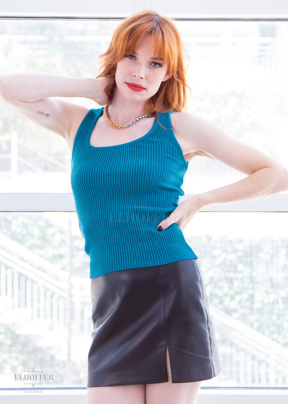 Chloe, a fair skinned XS model with red hair and bangs, is wearing a pullover sleeveless ribbed sweater tank with an asymmetrical neckline in teal.