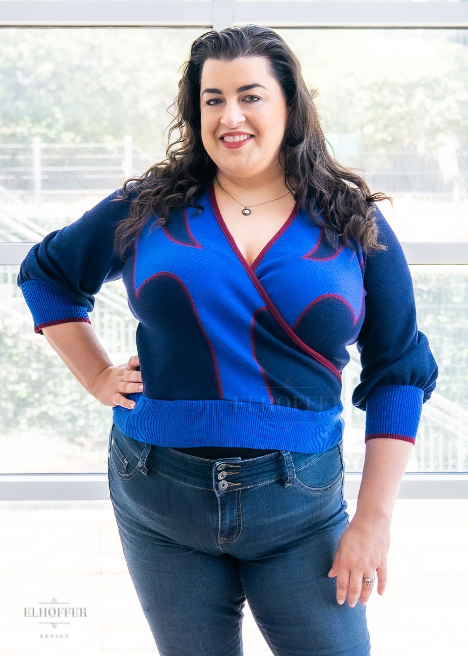 Heather, a size XL medium fair skinned model with long curly brown hair, wearing a crossover knit crop top. The base is a dark blue, details are bright blue with a red outline and form almost a t shape in the middle of the crop. The cuffs and bottom are the same bright blue of the detail and the crossover is piped in the same red.