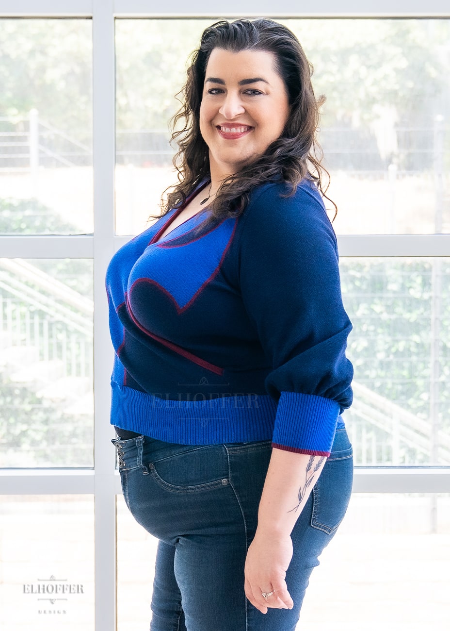 Heather, a size XL medium fair skinned model with long curly brown hair, wearing a crossover knit crop top. The base is a dark blue, details are bright blue with a red outline and form almost a t shape in the middle of the crop. The cuffs and bottom are the same bright blue of the detail and the crossover is piped in the same red. She stands sideways giving a better look at the three quarter sleeves.