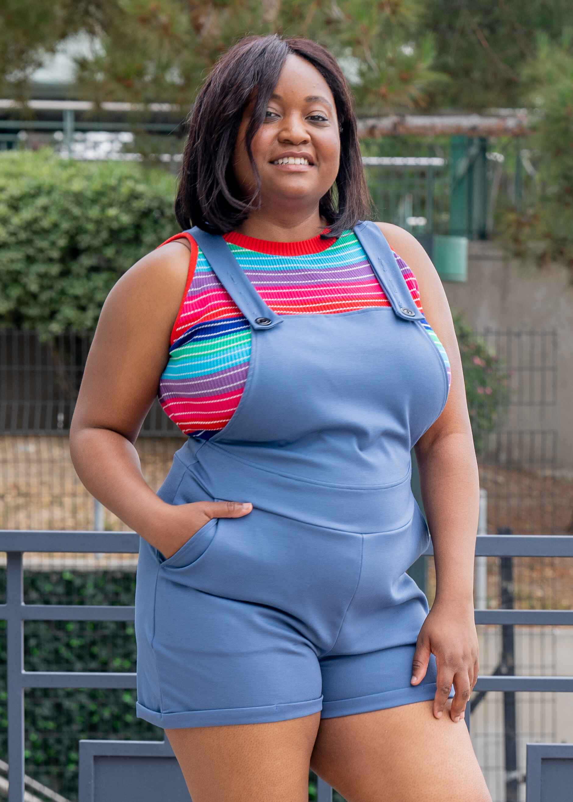 Maydelle, a medium dark skinned XL model with short dark brown hair, is smiling while wearing light denim blue short overalls. The overalls have side pockets and multiple buttons on each strap to make it easy to adjust. She paired the overalls with a rainbow striped sleeveless knit crop top.