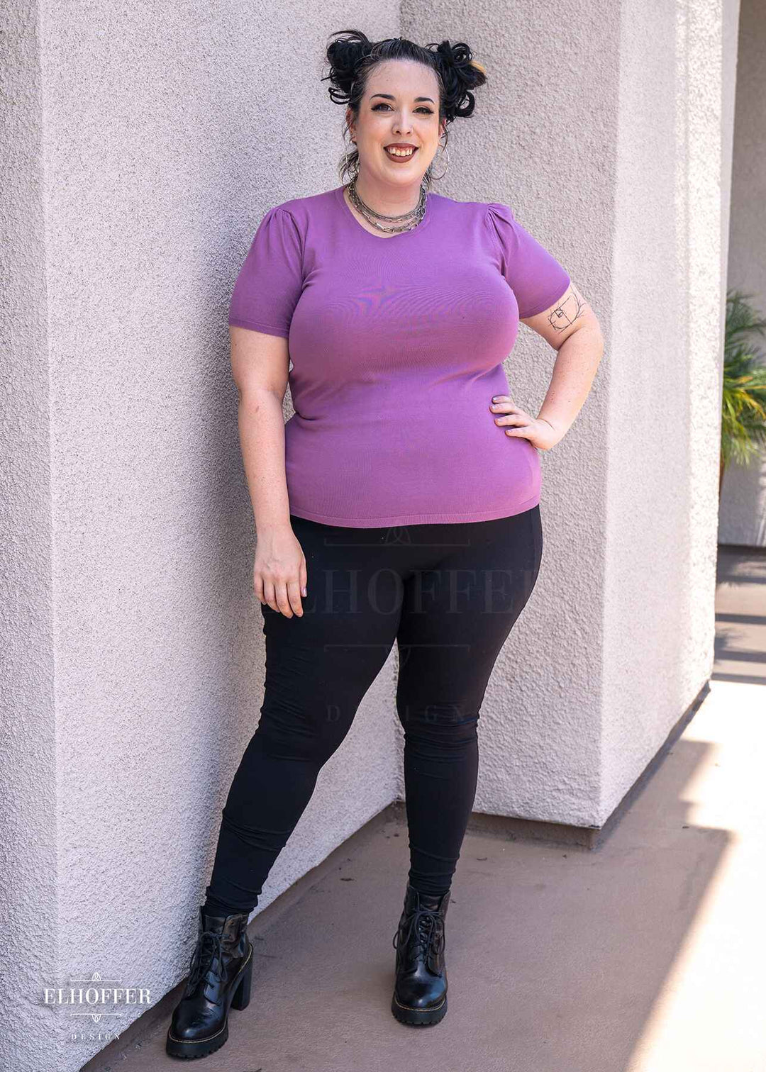 Katie Lynn, a fair skinned 2xl model with dark hair in space buns, is smiling while wearing a short sleeve light weight soft purple knit top. The top hits about mid hip in length and the sleeves have pleated gathering at the shoulders.