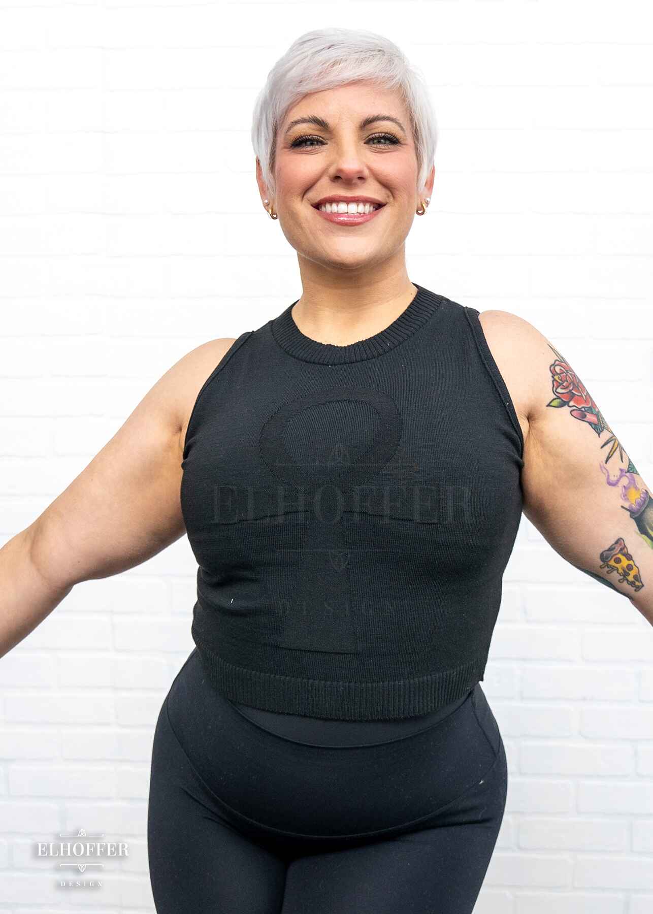 Whitney, a sun kissed skin L model with a platinum blonde pixie cut, is smiling while wearing a sleeveless knit crop top with a large subtle ankh design on the front.