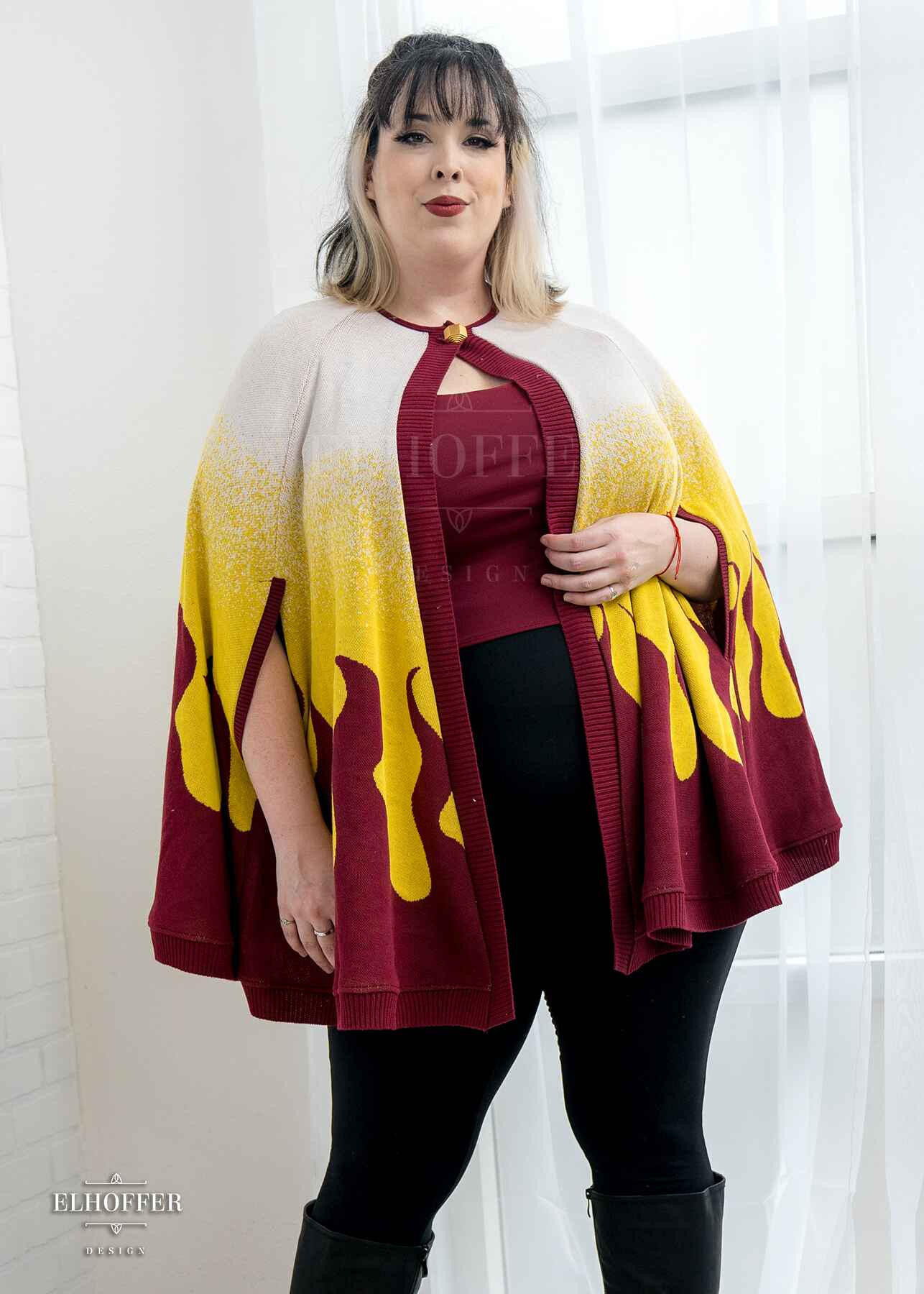 Katie Lynn, a fair skinned 2xl model with short black and blonde hair with bangs, is wearing a below hip length knit cape.  The cape is a gradient of color from white at the shoulders, to yellow, to a red fire design at the bottom, it also has a button closer at the neck, and armholes in the side seam. She paired the cape with a burgundy red scoop neck crop top and black leggings. 