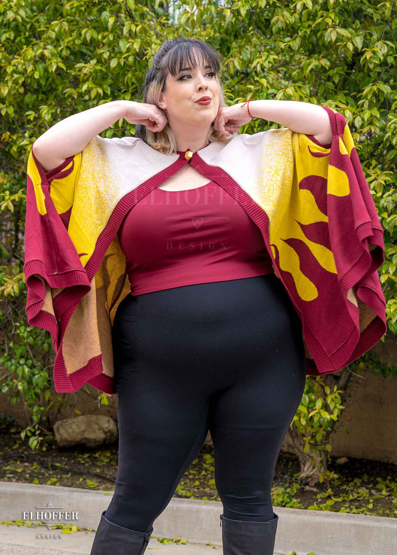 Katie Lynn, a fair skinned 2xl model with short black and blonde hair with bangs, is wearing a below hip length knit cape.  The cape is a gradient of color from white at the shoulders, to yellow, to a red fire design at the bottom, it also has a button closer at the neck, and armholes in the side seam. She paired the cape with a burgundy red scoop neck crop top and black leggings.