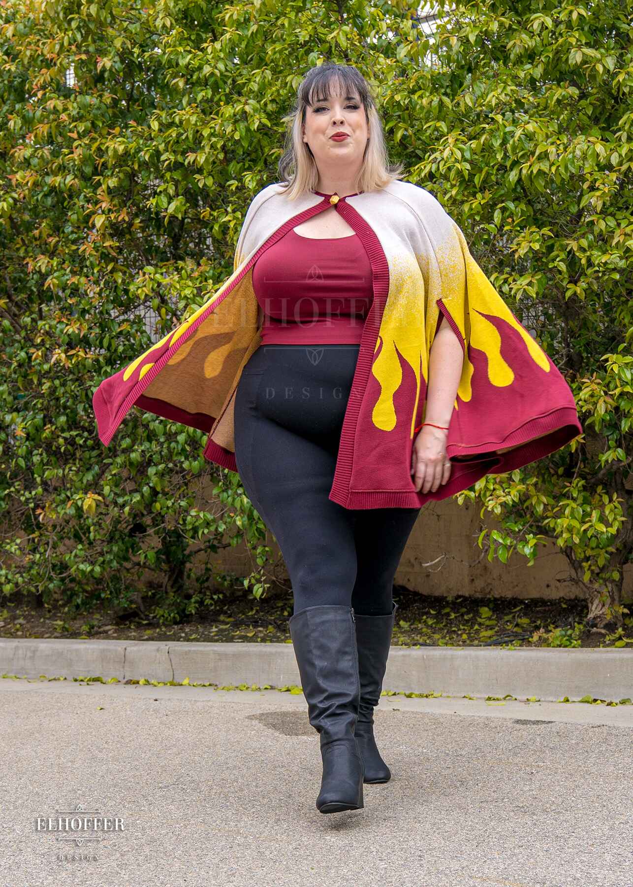 Katie Lynn, a fair skinned 2xl model with short black and blonde hair with bangs, is wearing a below hip length knit cape.  The cape is a gradient of color from white at the shoulders, to yellow, to a red fire design at the bottom, it also has a button closer at the neck, and armholes in the side seam. She paired the cape with a burgundy red scoop neck crop top and black leggings.