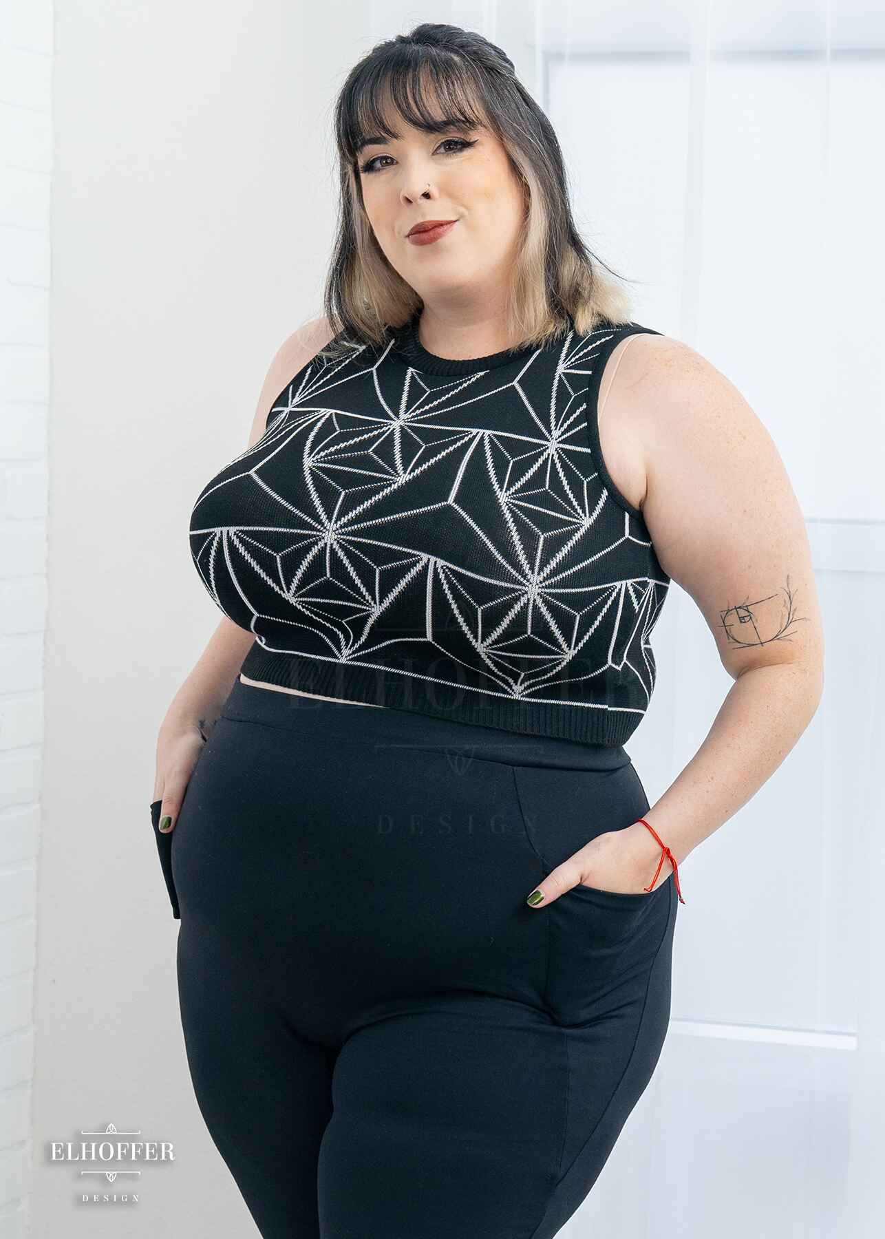 Katie Lynn, a fair skinned 2xl model with short black and white hair with bangs, is wearing a pullover cropped sleeveless sweater.  The main body of the sweater is black and has a white geometric 3d triangle design. She paired the crop top with black leggings.