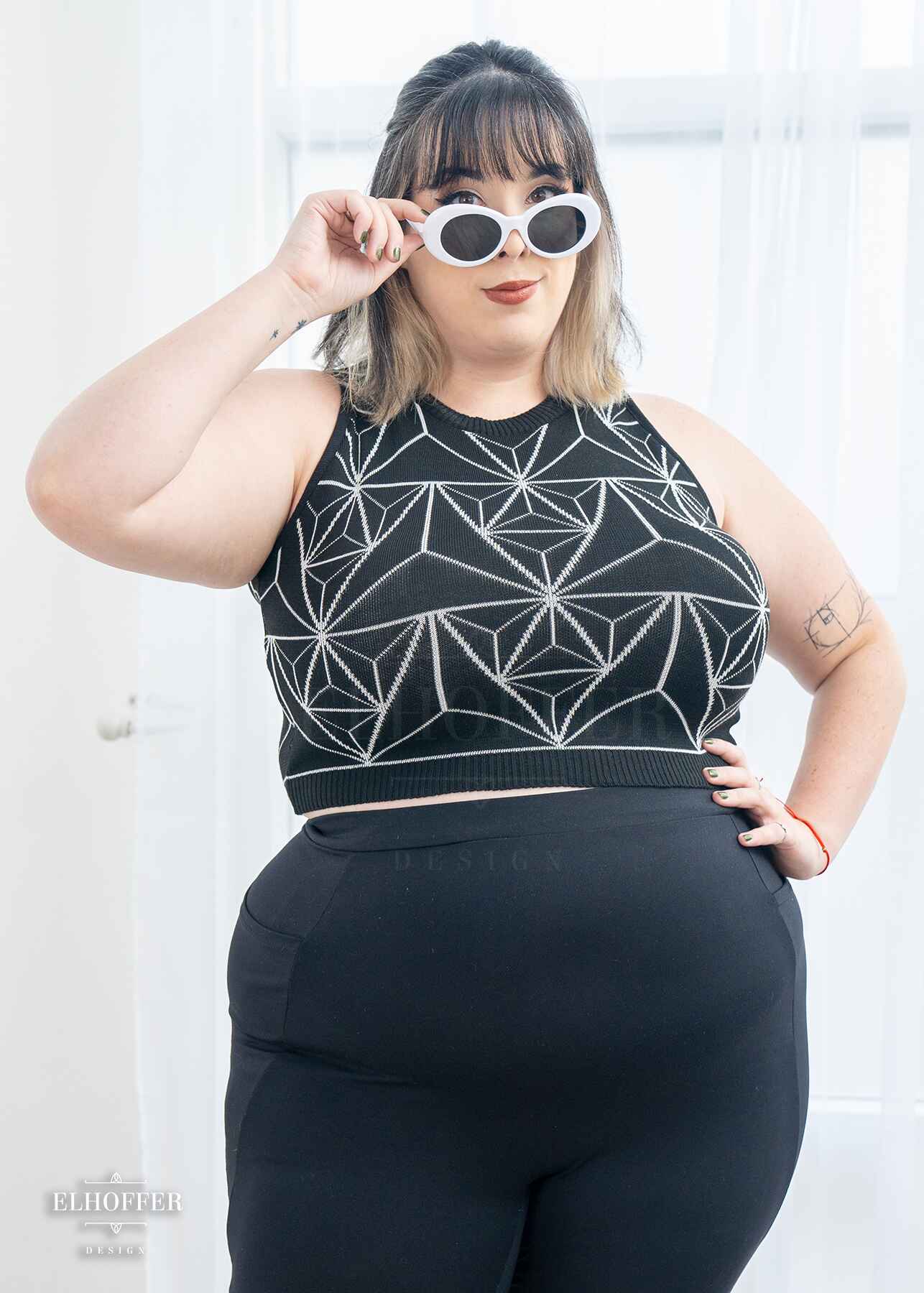 Katie Lynn, a fair skinned 2xl model with short black and white hair with bangs, is wearing a pullover cropped sleeveless sweater.  The main body of the sweater is black and has a white geometric 3d triangle design. She paired the crop top with black leggings.