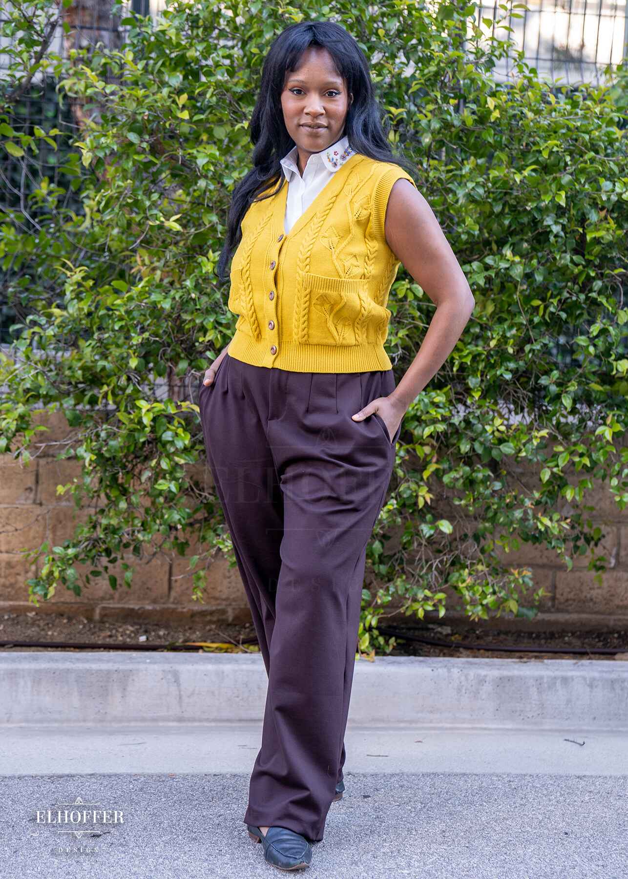 Lynsi, a medium dark skinned model with long black hair, is wearing chocolate brown wide leg trousers. The trousers feature a front zipper and button closure, pleated front, side pockets, and elastic in the back of the waistband for adjustability and cinch. She paired the trousers with a yellow knit vest with a leafy vine and cable knit design.
