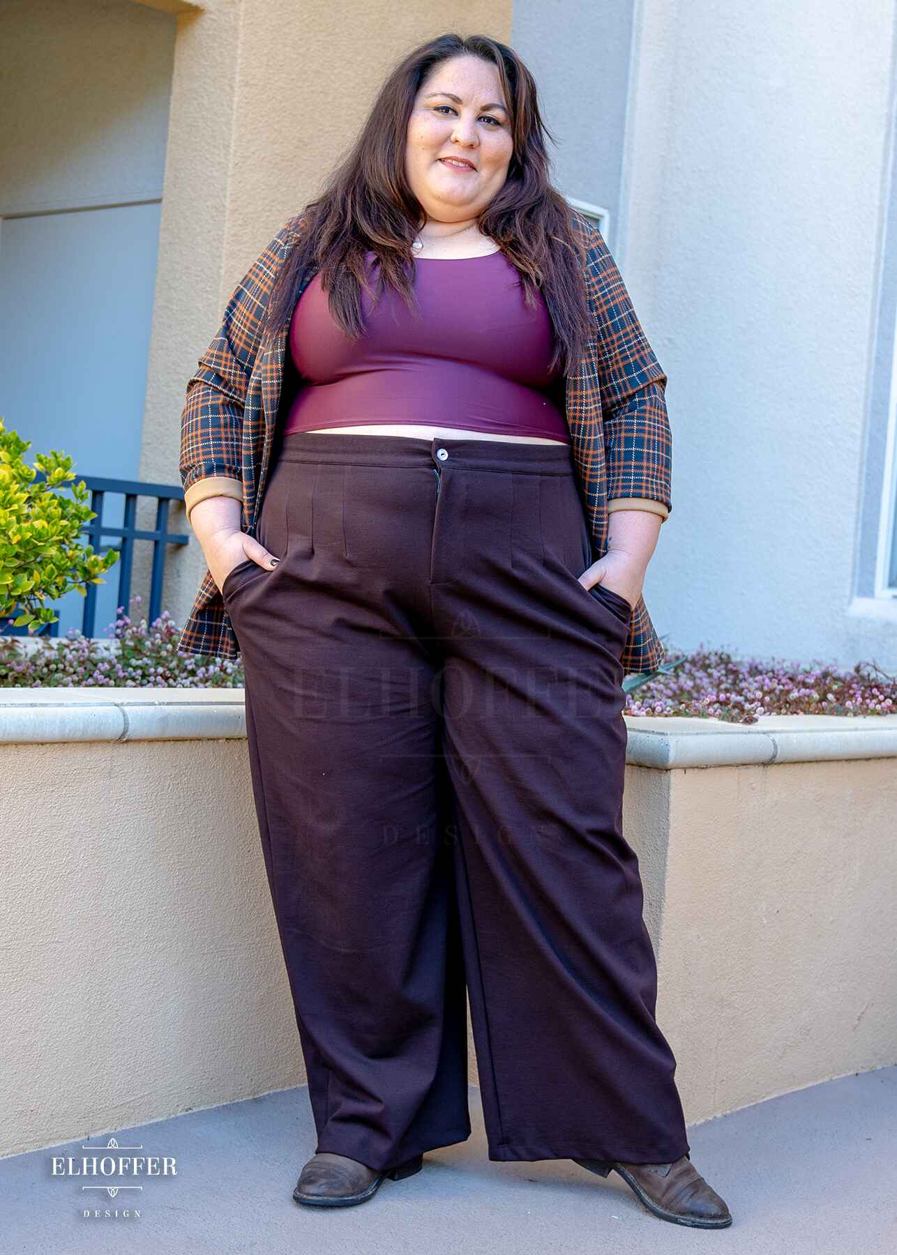 Alysia, a sun kissed skin 2xl model with long dark brown hair, is wearing chocolate brown wide leg trousers.  The trousers feature a front zipper and button closure, pleated front, side pockets, and elastic in the back of the waistband for adjustability and cinch. She paired the trousers with a dark purplish red scoop neck sleeveless crop top.