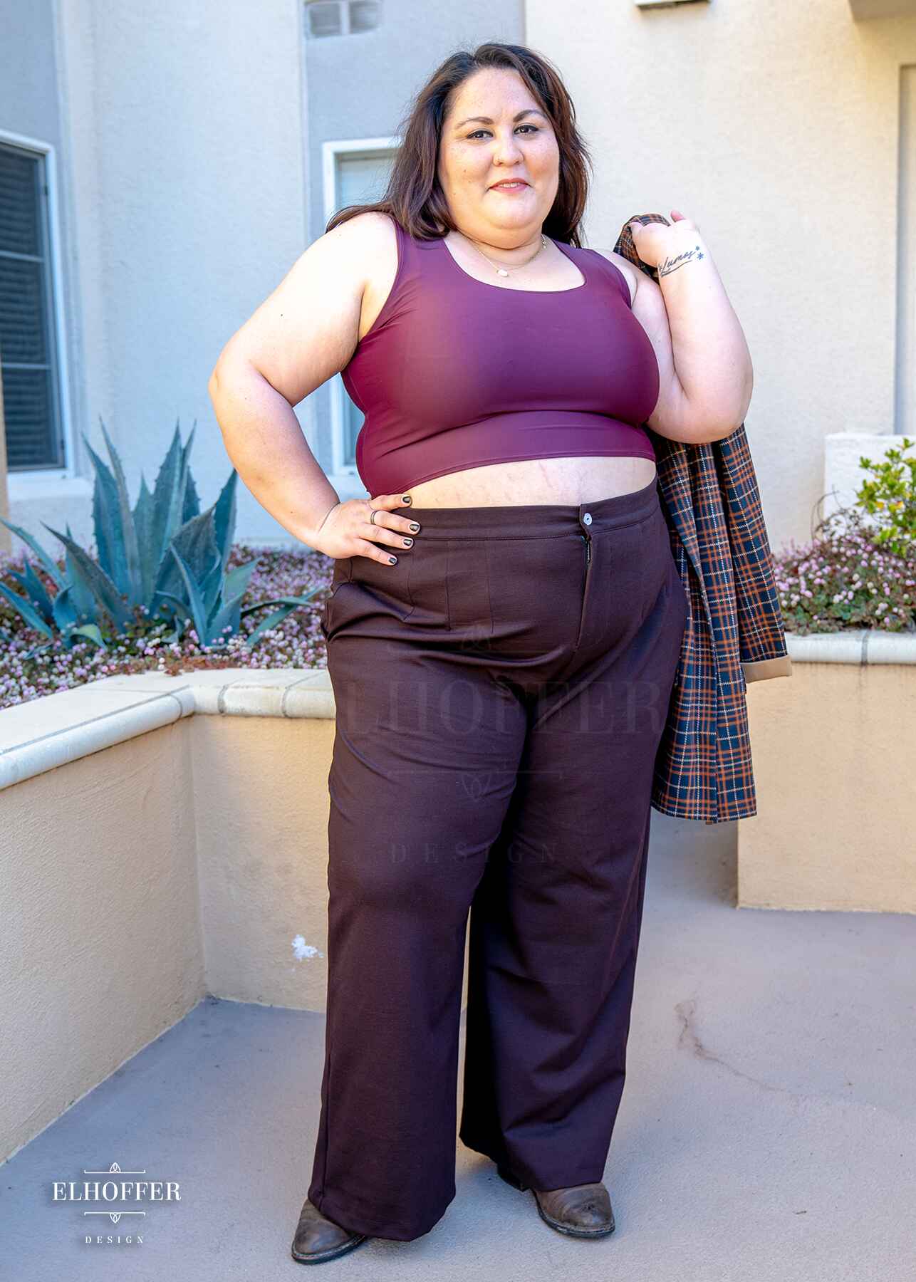 Alysia, a sun kissed skin 2xl model with long dark brown hair, is wearing chocolate brown wide leg trousers.  The trousers feature a front zipper and button closure, pleated front, side pockets, and elastic in the back of the waistband for adjustability and cinch. She paired the trousers with a dark purplish red scoop neck sleeveless crop top.