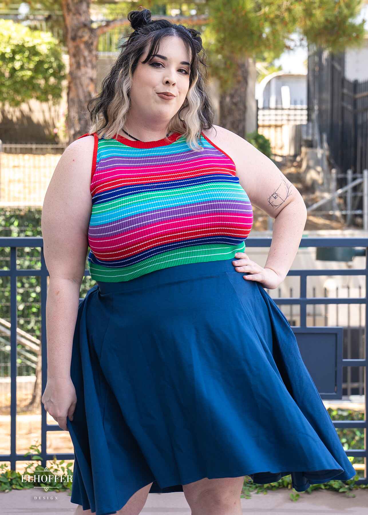 Katie Lynn, a fair skinned 2xl model with shoulder length black and blonde hair is wearing a sleeveless striped knit crop top. The crop top has thicker stripes of green, blue, red, pink, purple, and teal with pinstripe white lines in each colored stripe. She paired the crop top with a knee length denim blue high low skirt. Shirt is also available in a longer version.
