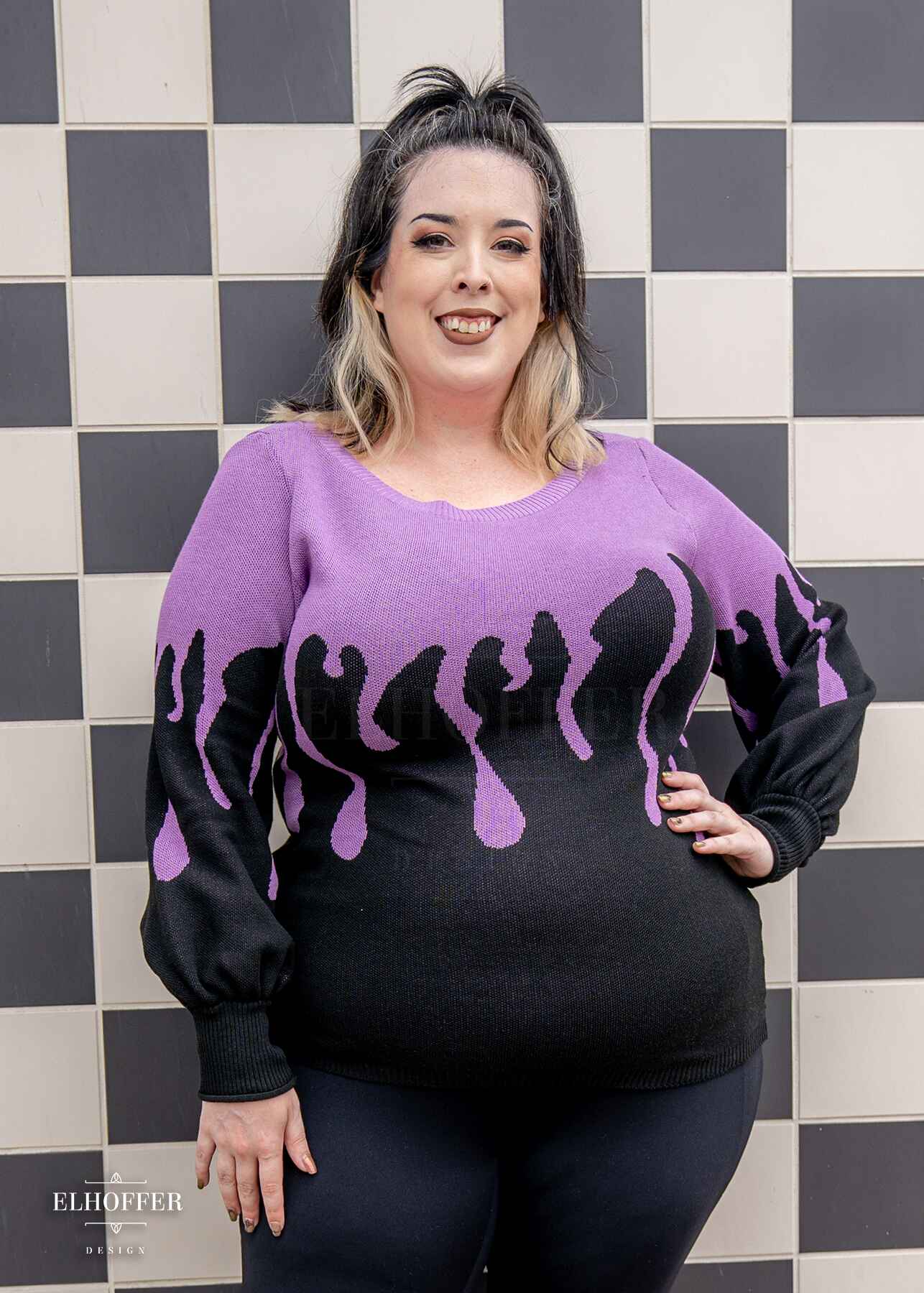 Katie Lynn, a fair skinned 2xl model with shoulder length wavy black and white hair, is smiling while wearing an oversize sweater with a lavender purple drip design that looks like it's oozing down onto a black sweater that has long billowing sleeves with thumbholes.