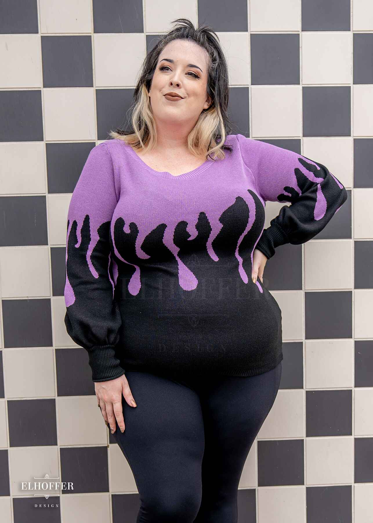Katie Lynn, a fair skinned 2xl model with shoulder length wavy black and white hair, is wearing an oversize sweater with a lavender purple drip design that looks like it's oozing down onto a black sweater that has long billowing sleeves with thumbholes.