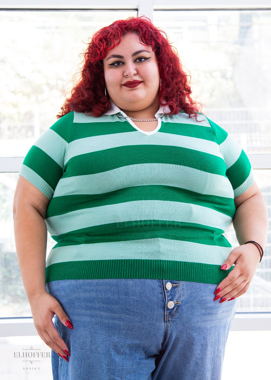 Victoria, an olive skinned 4XL model with bright red curly hair, is wearing a pullover fitted lightweight sweater with short sleeves and split neck with a small white collar. It's dark green with lighter green thick stripes running horizontally.