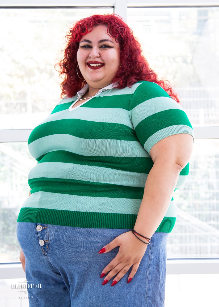 Victoria, an olive skinned 4XL model with bright red curly hair, is wearing a pullover fitted lightweight sweater with short sleeves and split neck with a small white collar. It's dark green with lighter green thick stripes running horizontally.