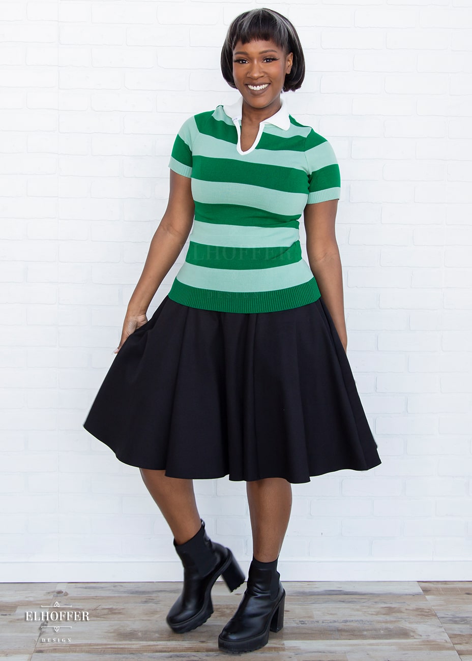 Lynsi, a medium dark skinned model with a short dark brown and white bob, is wearing a pullover fitted lightweight sweater with short sleeves and split neck with a small white collar. It's dark green with lighter green thick stripes running horizontally.