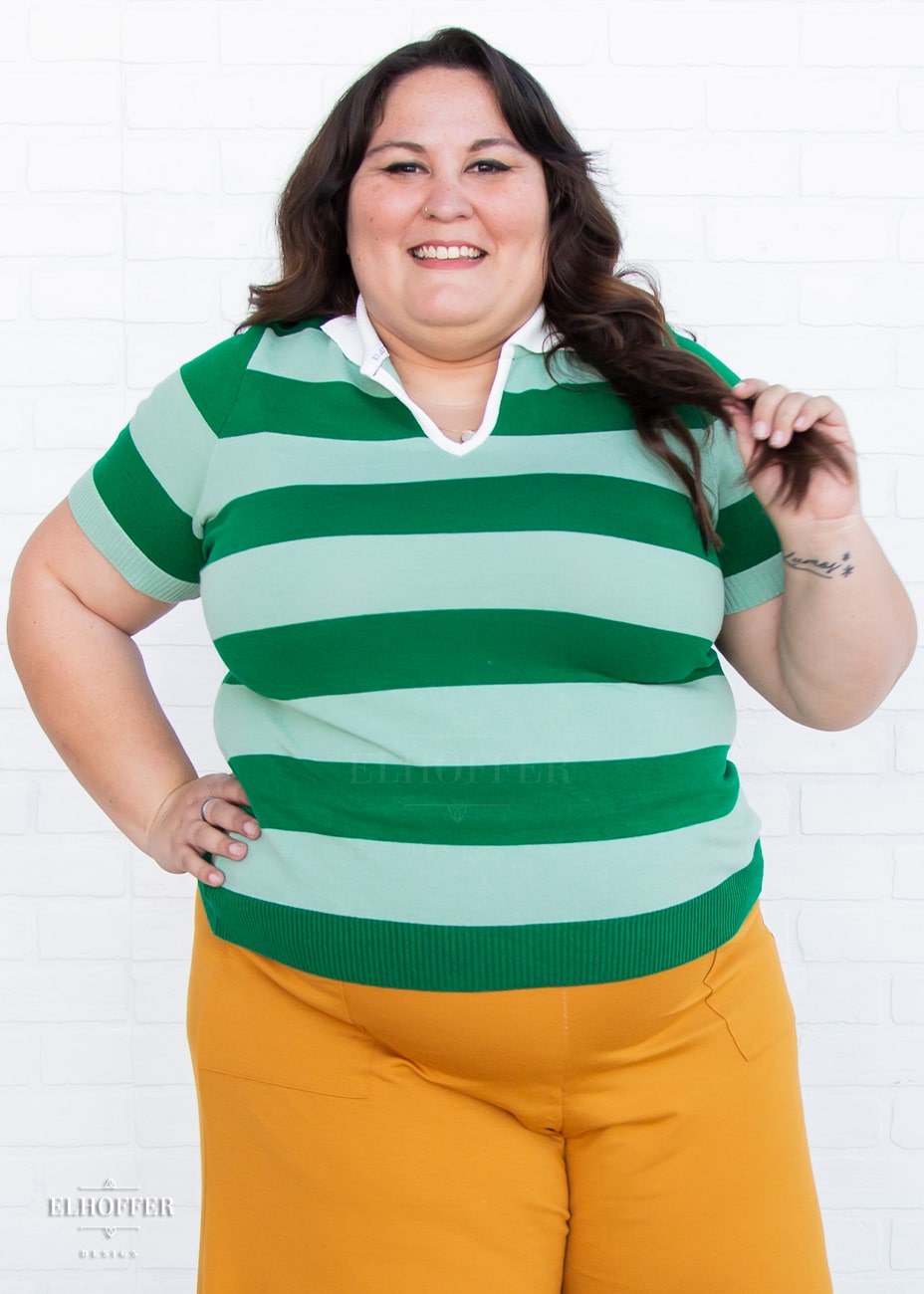 Alysia, a size 2XL model with fair skin and long dark brown hair, is wearing a pullover fitted lightweight sweater with short sleeves and split neck with a small white collar. It's dark green with lighter green thick stripes running horizontally.