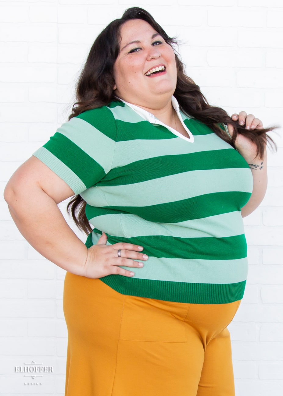 Alysia, a size 2XL model with fair skin and long dark brown hair, is wearing a pullover fitted lightweight sweater with short sleeves and split neck with a small white collar. It's dark green with lighter green thick stripes running horizontally.