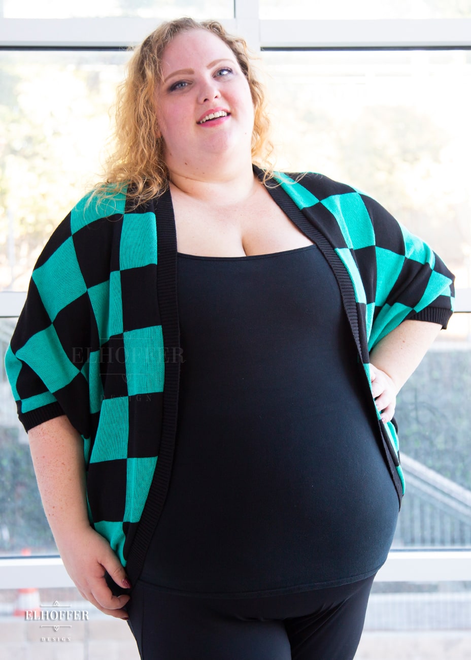 Bee, a fair skinned 3xl model with shoulder length curly blonde hair, is wearing a XL-3XL sample of a shrug with a black and green chessboard pattern. The shrug featured 3/4 sleeves and black ribbing along edges and cuffs.