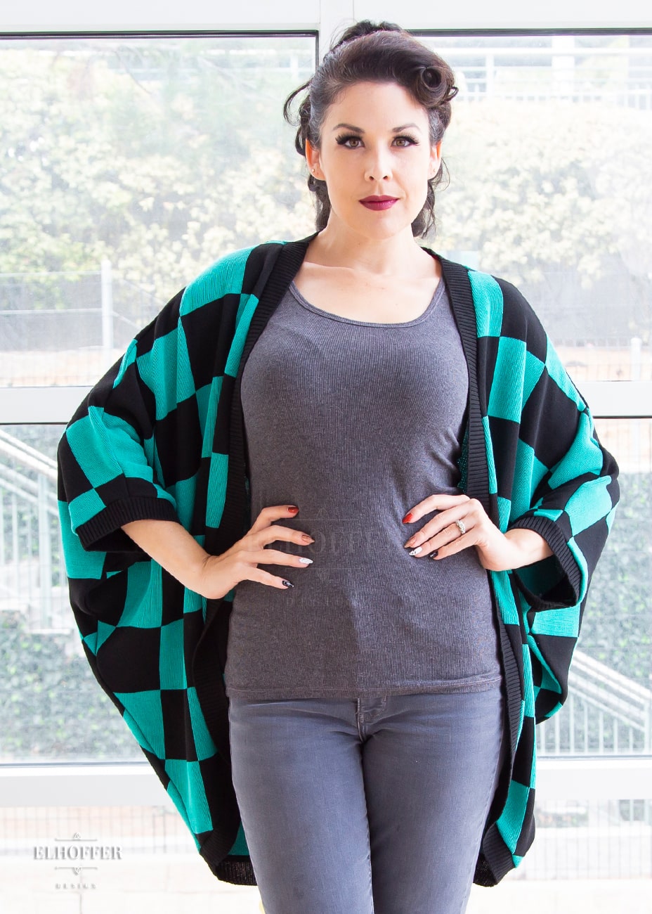 Kit, a fair skinned S model with long dark brown hair pulled back in a pony tail, is wearing a XL-3XL sample of a shrug with a black and green chessboard pattern. The shrug featured 3/4 sleeves and black ribbing along edges and cuffs. The XL-3XL sample is oversized on her, she would order the XS-L for a more fitted look.