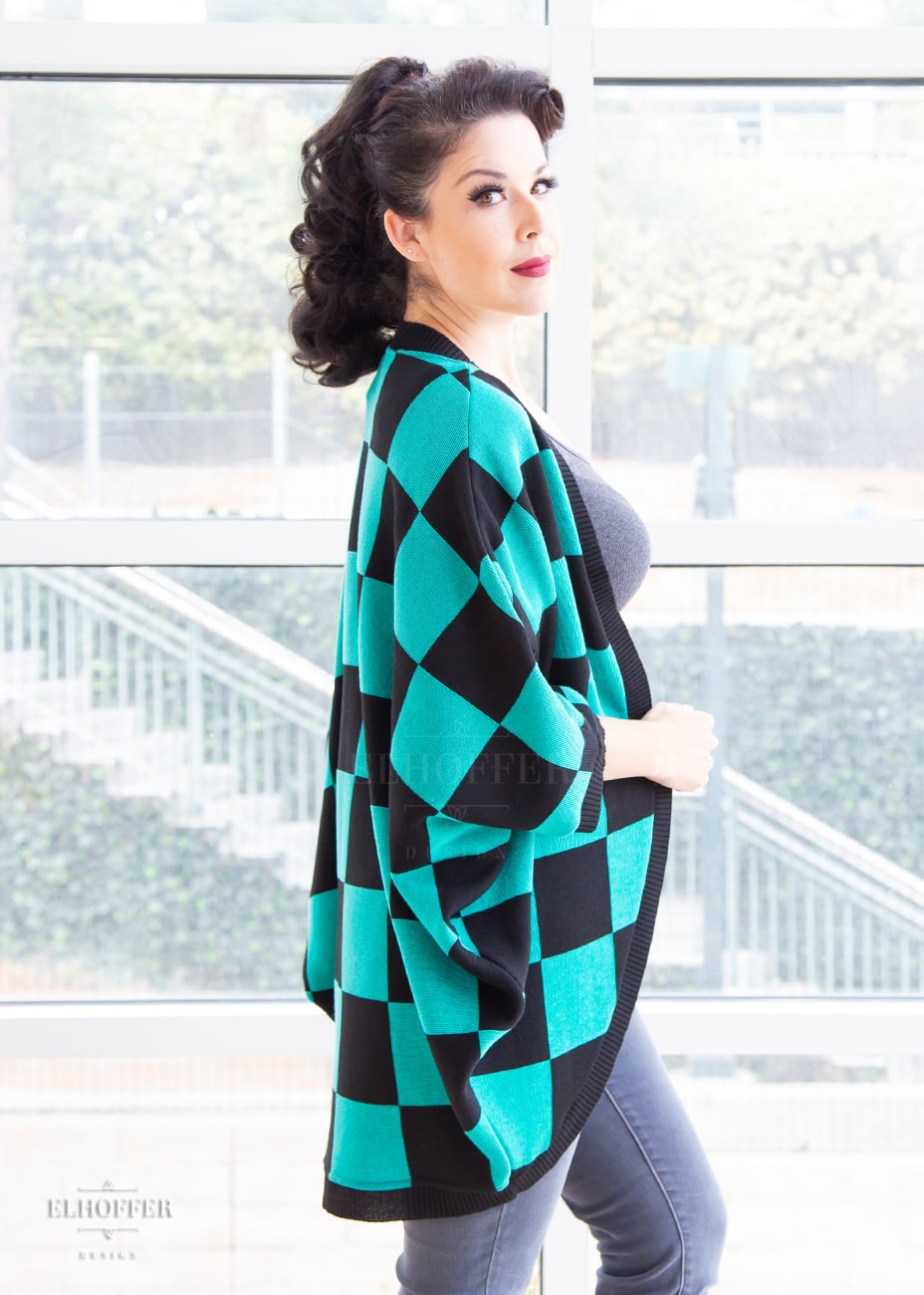 A side view of Kit, a fair skinned S model with long dark brown hair pulled back in a pony tail, wearing a XL-3XL sample of a shrug with a black and green chessboard pattern. The shrug featured 3/4 sleeves and black ribbing along edges and cuffs. The XL-3XL sample is oversized on her, she would order the XS-L for a more fitted look.