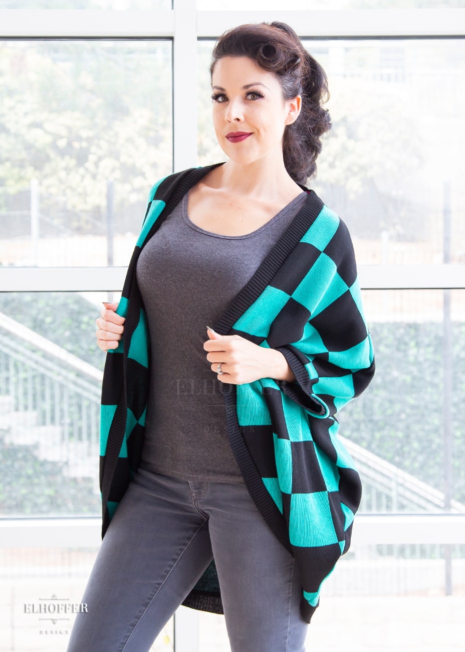 Kit, a fair skinned S model with long dark brown hair pulled back in a pony tail, is wearing a XL-3XL sample of a shrug with a black and green chessboard pattern. The shrug featured 3/4 sleeves and black ribbing along edges and cuffs. The XL-3XL sample is oversized on her, she would order the XS-L for a more fitted look.