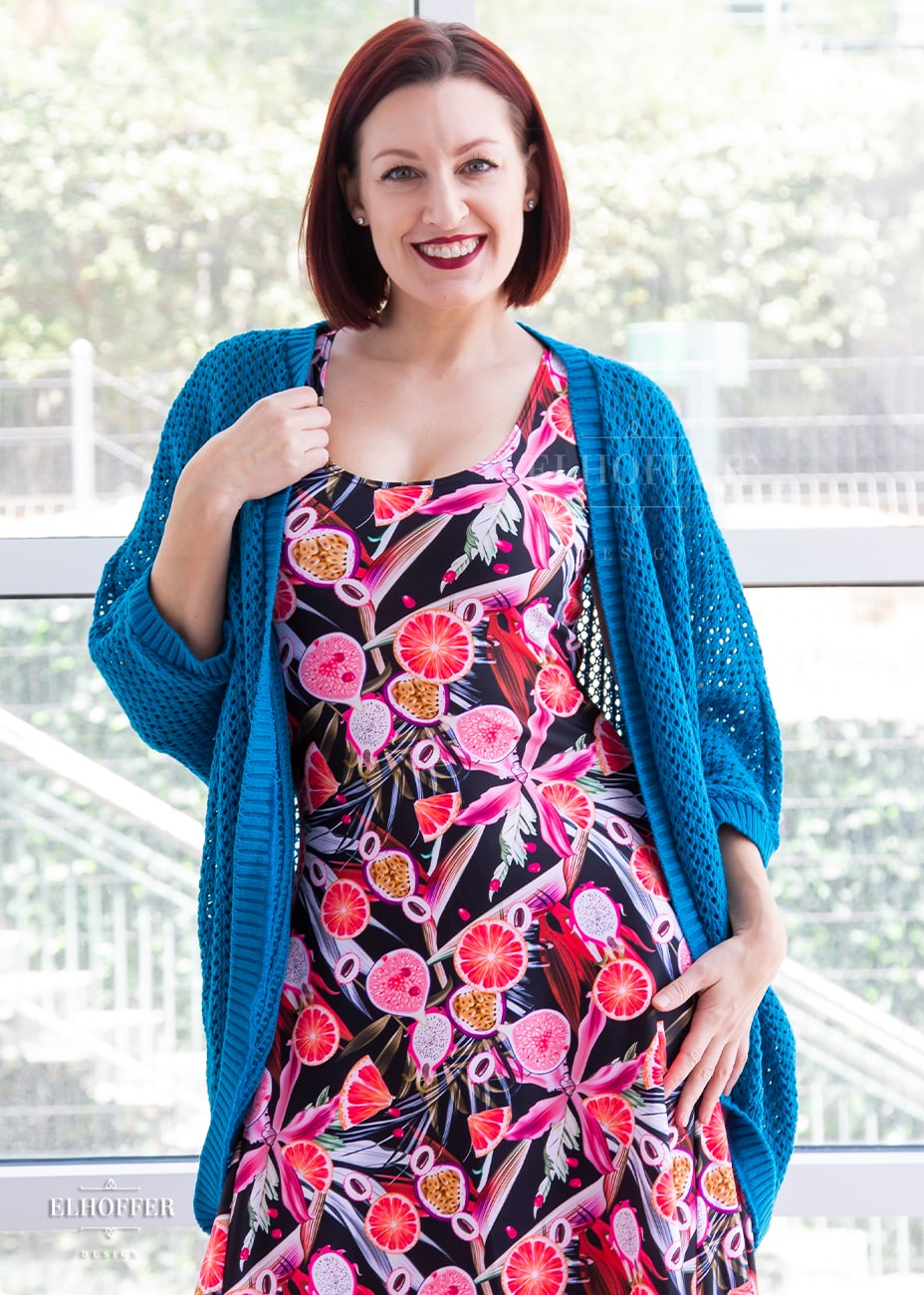 Natalie, a size small fair skinned model with short red hair, is wearing an open front dolman shrug with a loose knit in a medium blue.